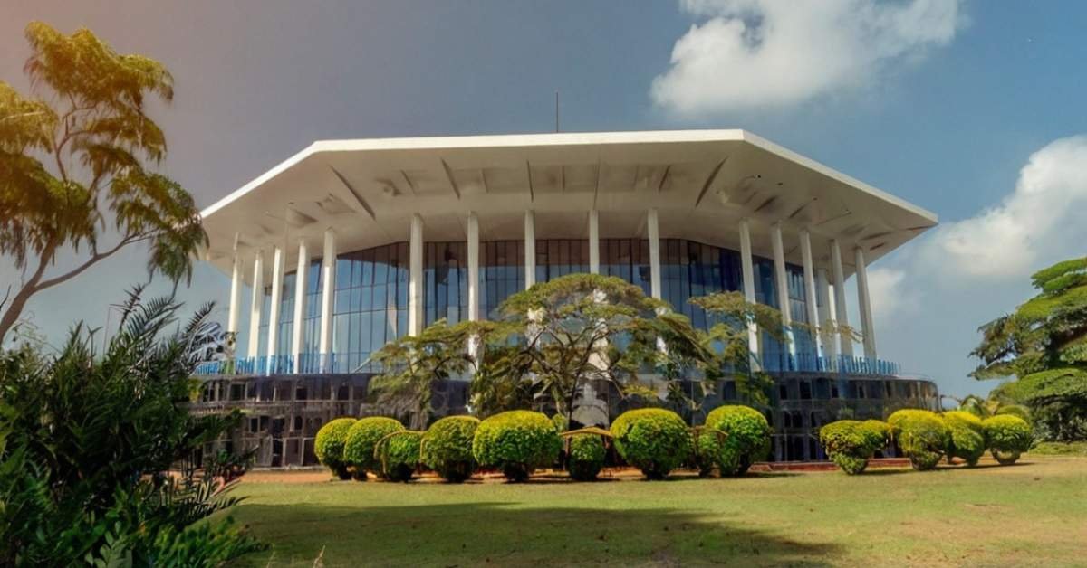 A large, white, modern building with a distinctive octagonal roof and numerous tall, slender white columns supporting it. The building features extensive glass windows reflecting the sky. It is surrounded by a well-maintained green lawn with various trees and manicured shrubs. The sky above is blue with scattered white clouds.