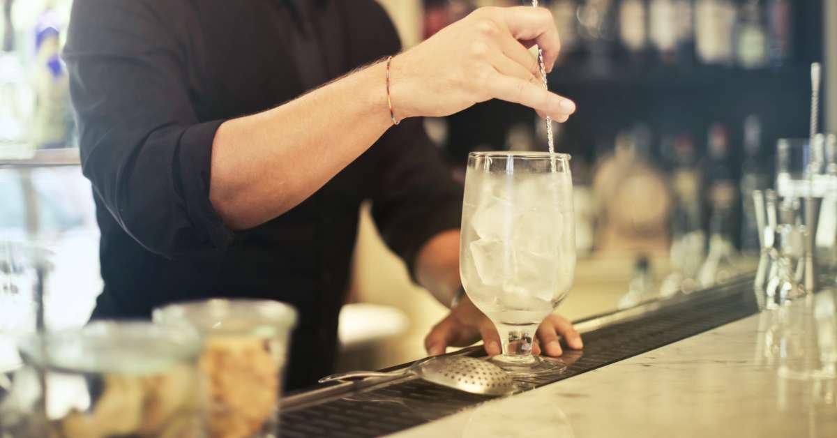 A bartender in a black shirt stirs ice in a tall glass on a bar counter.