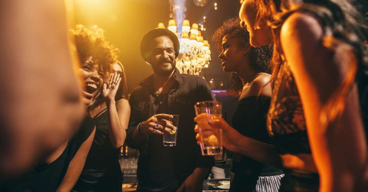 A diverse group of stylish young adults laughing and socializing over drinks in a vibrant, warmly lit nightclub with a chandelier in the background.